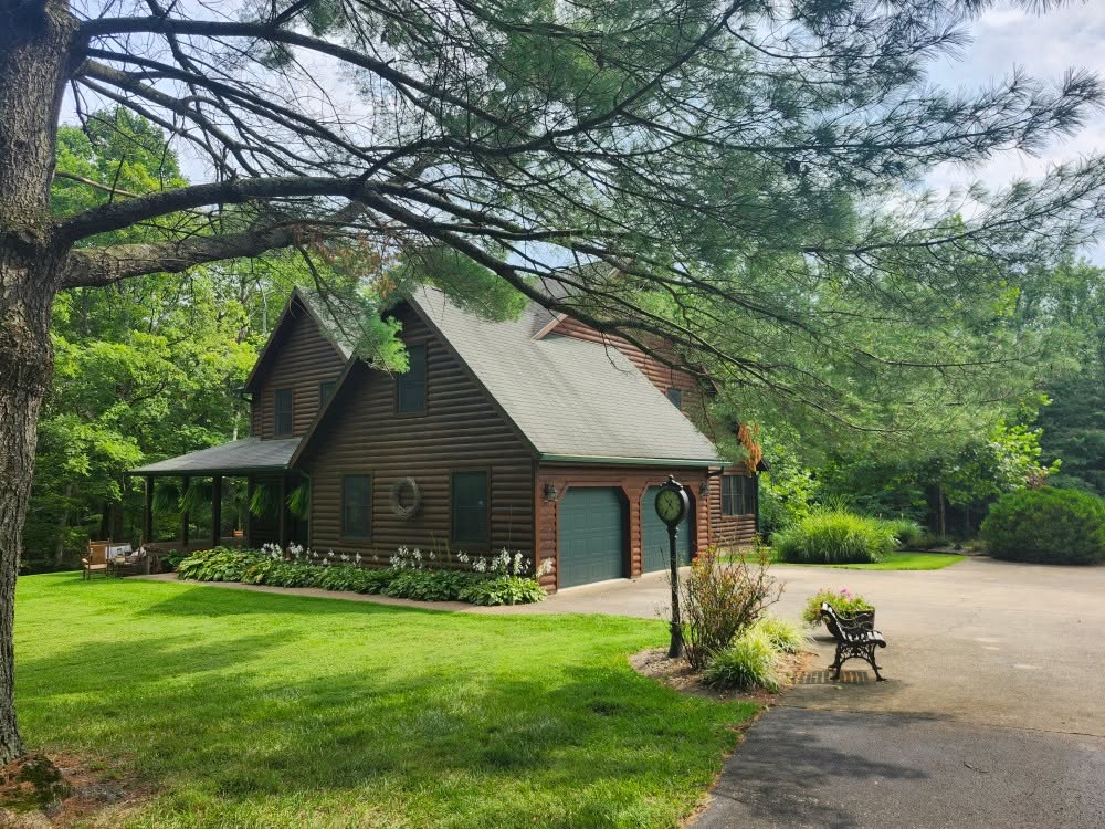 Log home with a fresh brown stain, green garage doors and a wraparound porch, set among tall trees