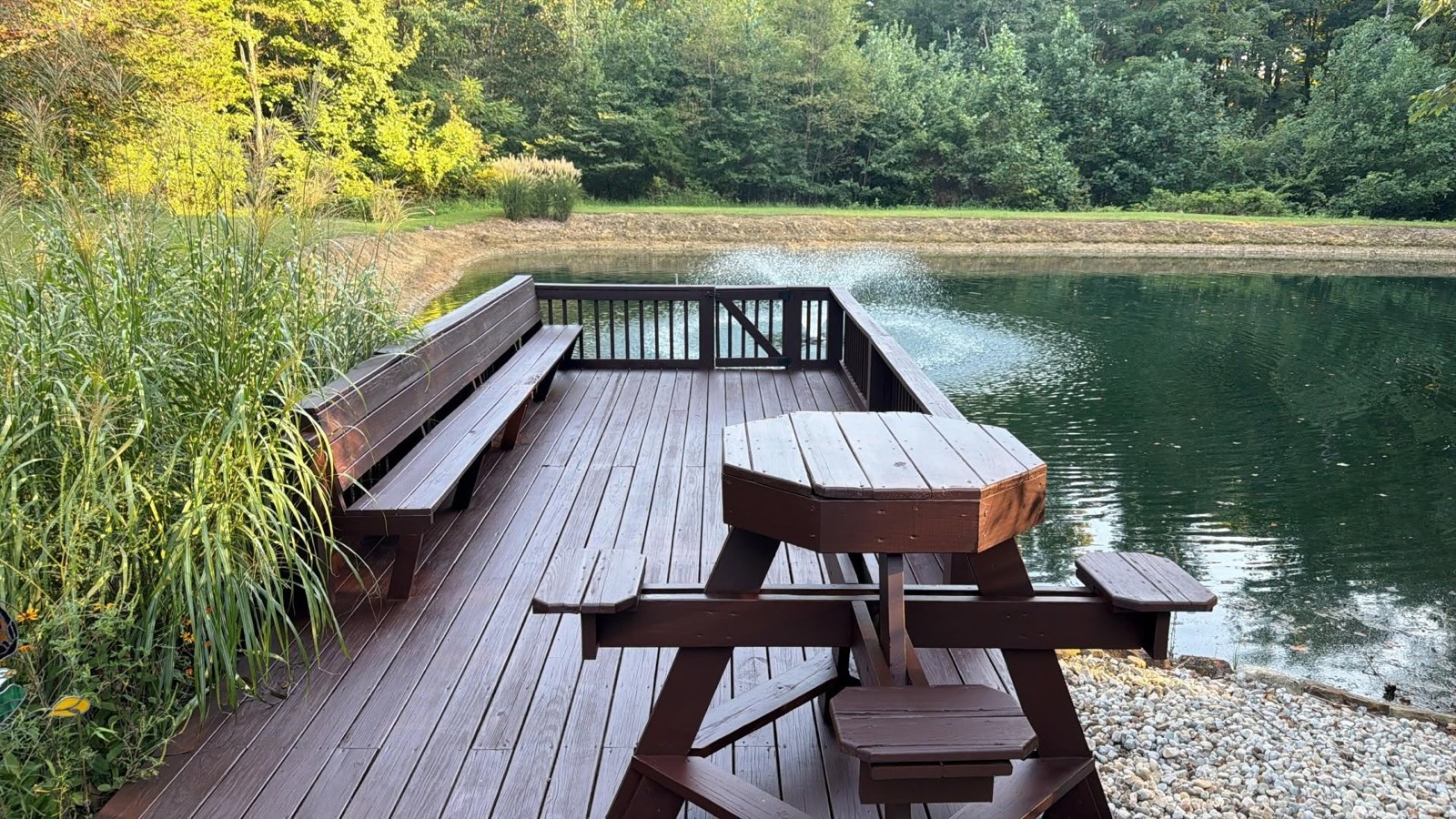 Stained wooden deck with built-in benches and a picnic table overlooking a pond with a fountain